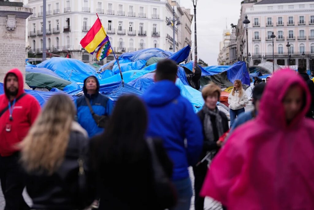 Transeúntes caminan frente a un campamento cubierto con lonas azules y banderas en la Puerta del Sol de Madrid.